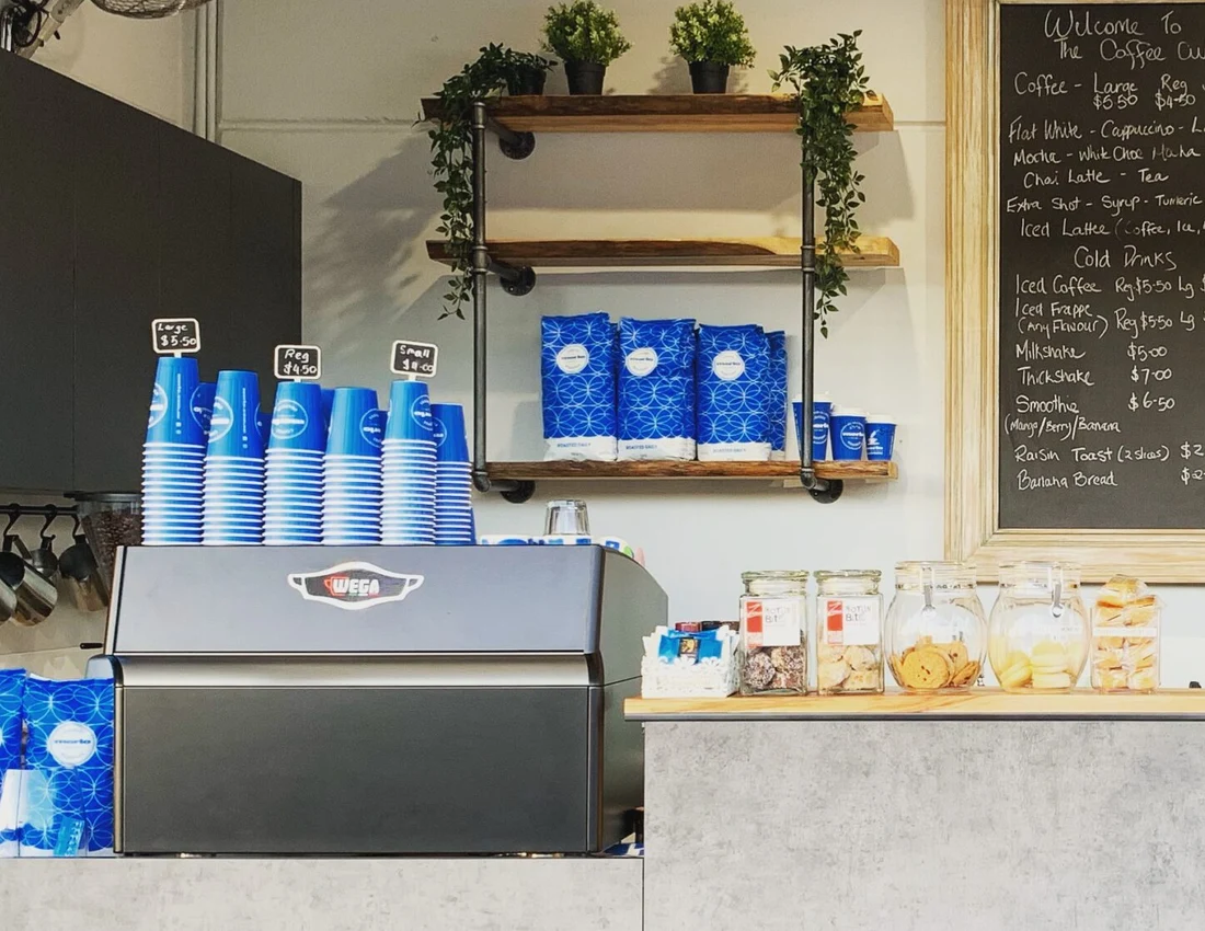 Blue Merlo cups and coffee beans at Coffee Cube’s espresso machine, surrounded by timber shelves and plants